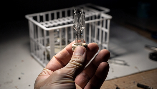 A bare projector lamp burner with twin quartz arc tubes held in gloved fingertips against a dark background