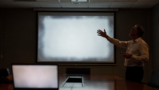 A man in a dim meeting room gesturing toward a projection screen showing a dim, washed-out image